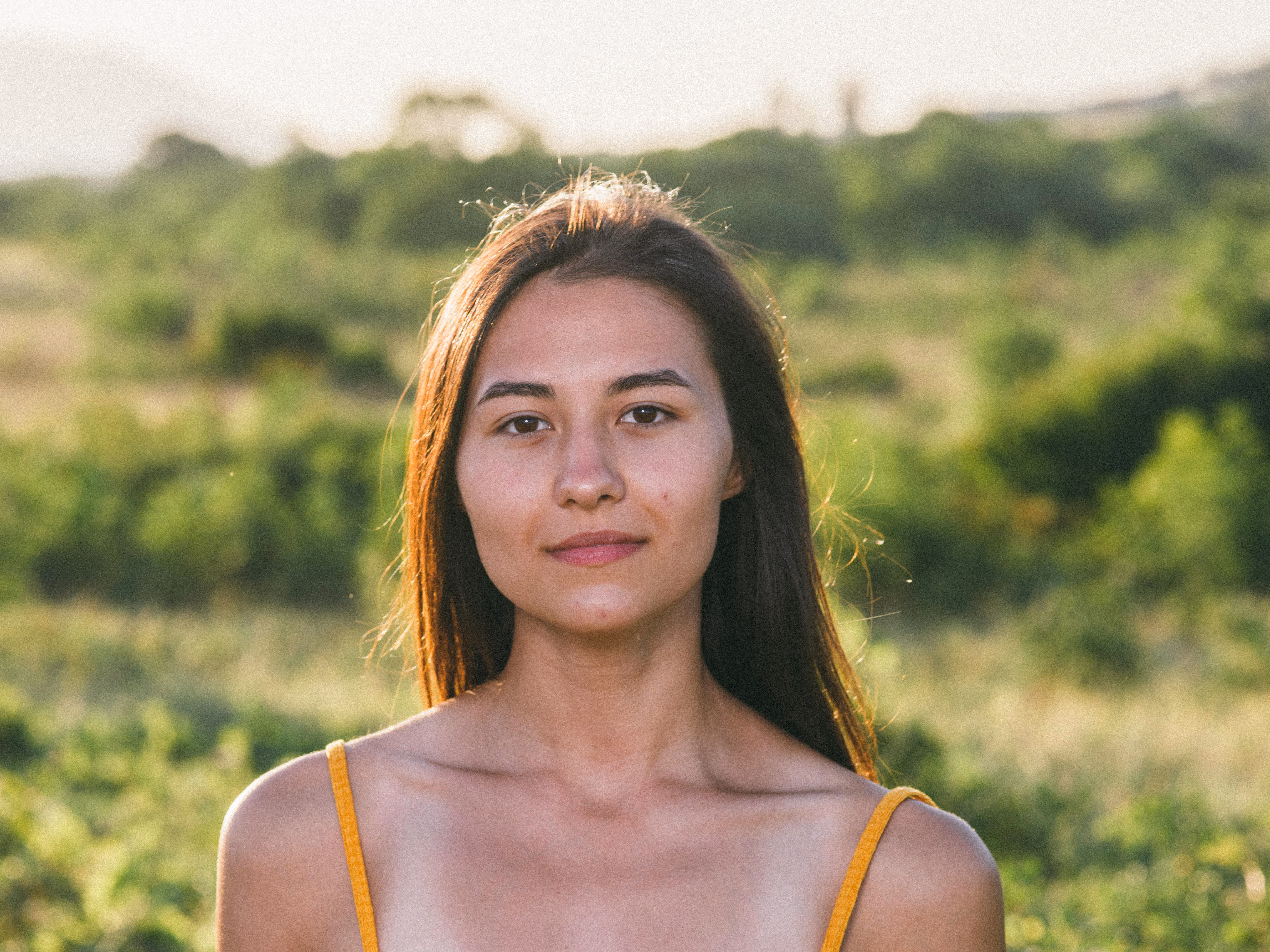 Junge Frau mit langen dunklen Haaren und gelbem ärmellosem Top in einer grünen Landschaft bei Sonnenlicht.